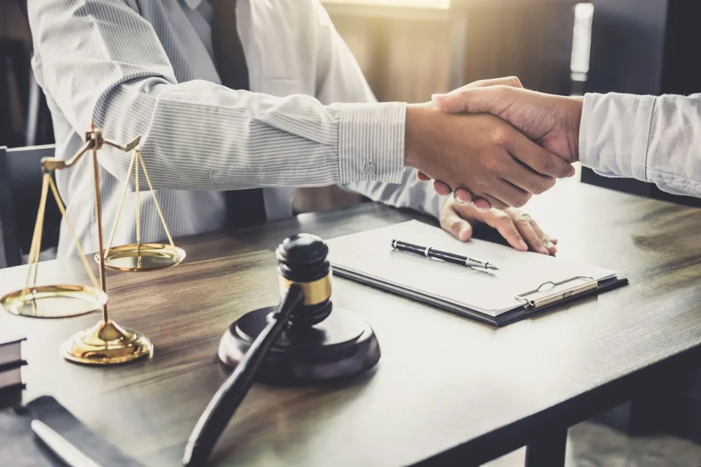 Two people shaking hands across a desk featuring a wooden gavel, scales, and a document with a pen, representing business litigation in Irvine.