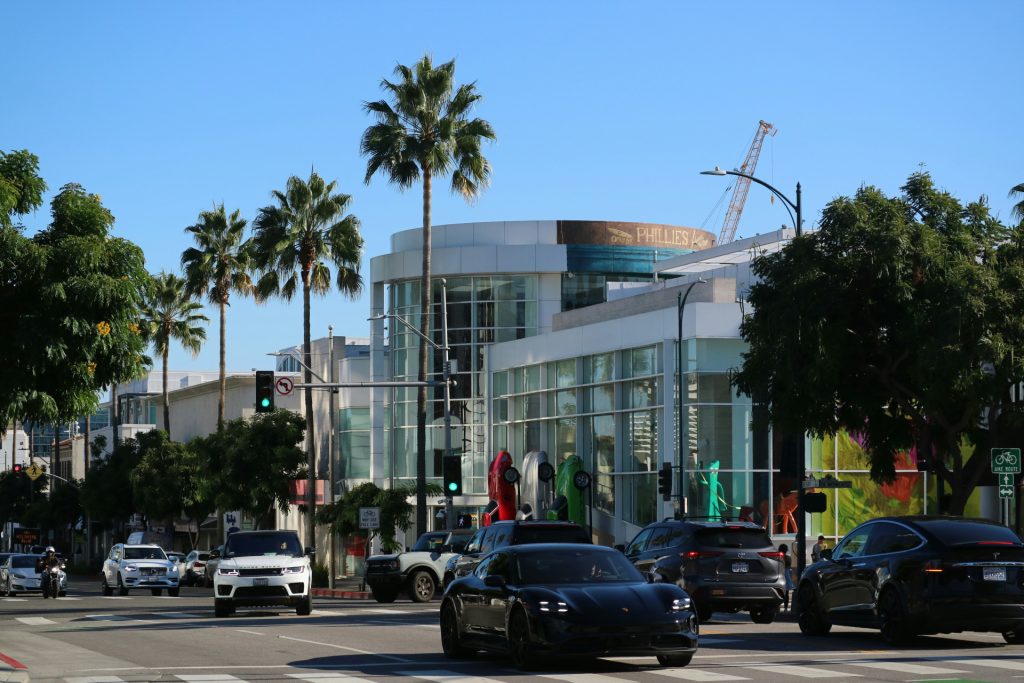 Traffic moves through a busy intersection in front of a modern glass building where a business lawyer in Beverly Hills might work, marked by a “Phillies” sign and palm trees.