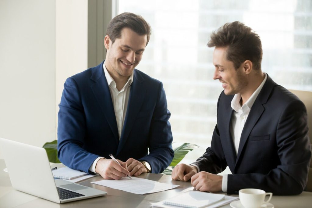 Two men in business lawyer suits sit at a desk, one signing a document while the other watches and smiles in West Los Angeles.