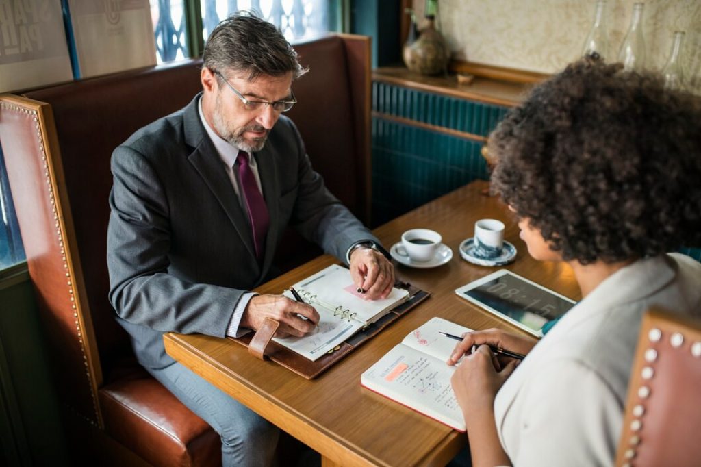 A business lawyer in Greater Los Angeles sits in a booth with a woman, both writing in their respective planners during a meeting.