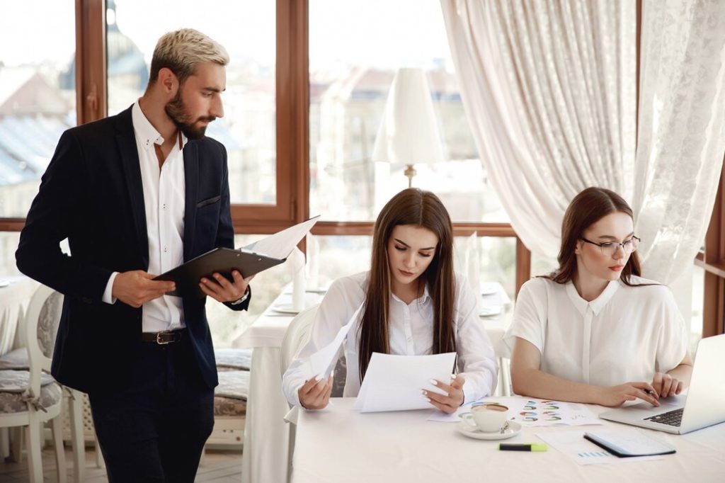 A business lawyer serving the South Bay stands to review documents while two women sit at a table working with papers and a laptop.