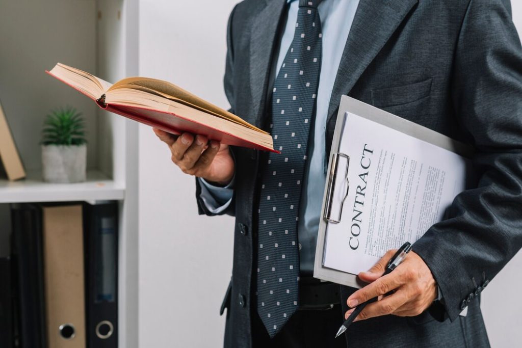 A business lawyer in Koreatown in a suit holds an open book and a clipboard labeled “CONTRACT” with a pen in hand.