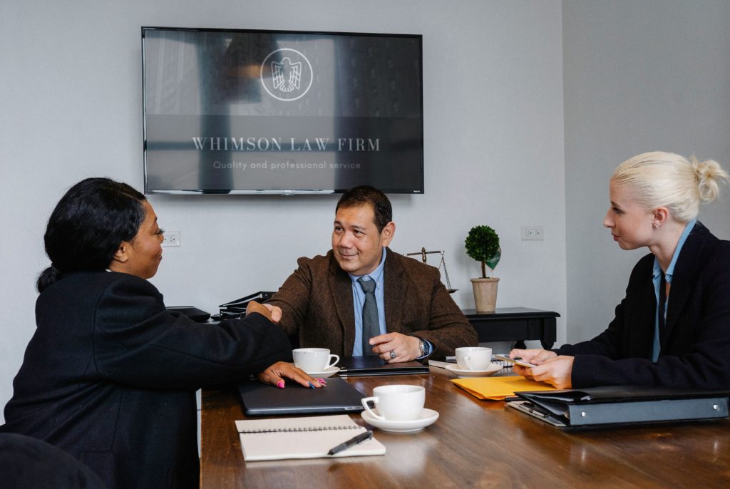Three business lawyers in Malibu shake hands across a wooden conference table in an office with a law firm logo displayed on a wall-mounted screen.