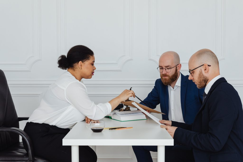 A business lawyer in Mid-City points to a document while consulting with two men in suits at a desk.