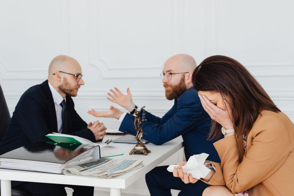 A woman holds a tissue to her face while sitting in front of a business lawyer in Mid-Wilshire and another man in suits who are having a discussion.