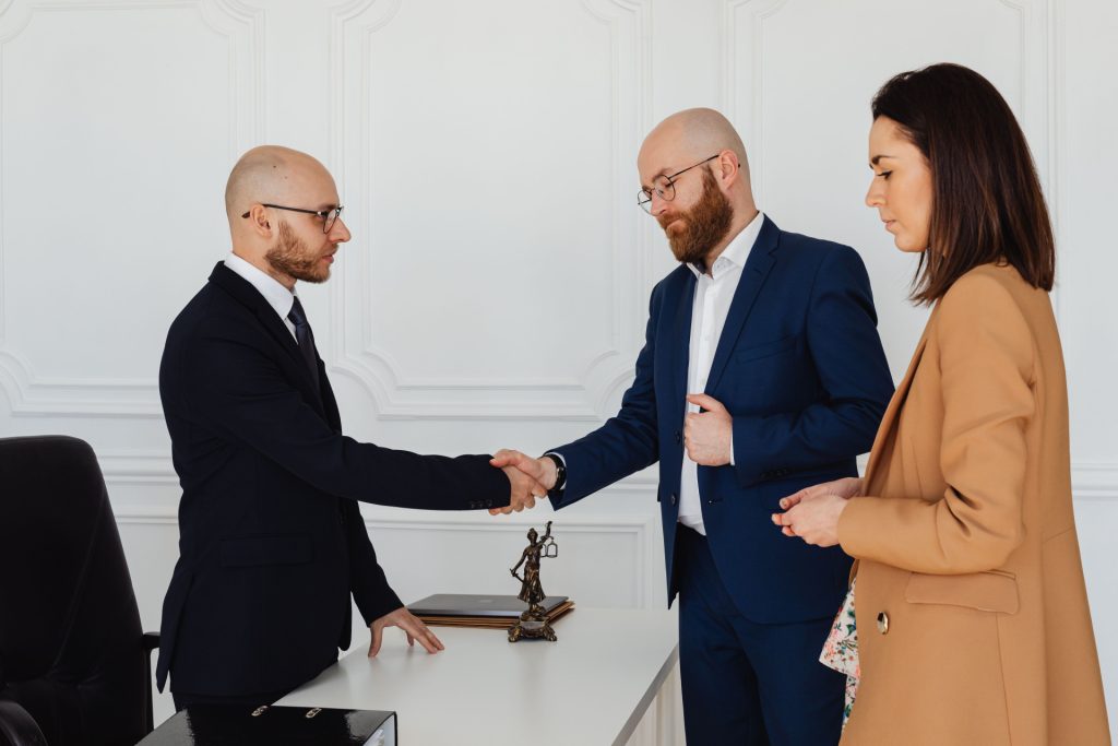 Two men in suits shaking hands across a desk, observed by a woman in a tan blazer, with a statue of Lady Justice on the desk of a business lawyer in Palos Verdes.