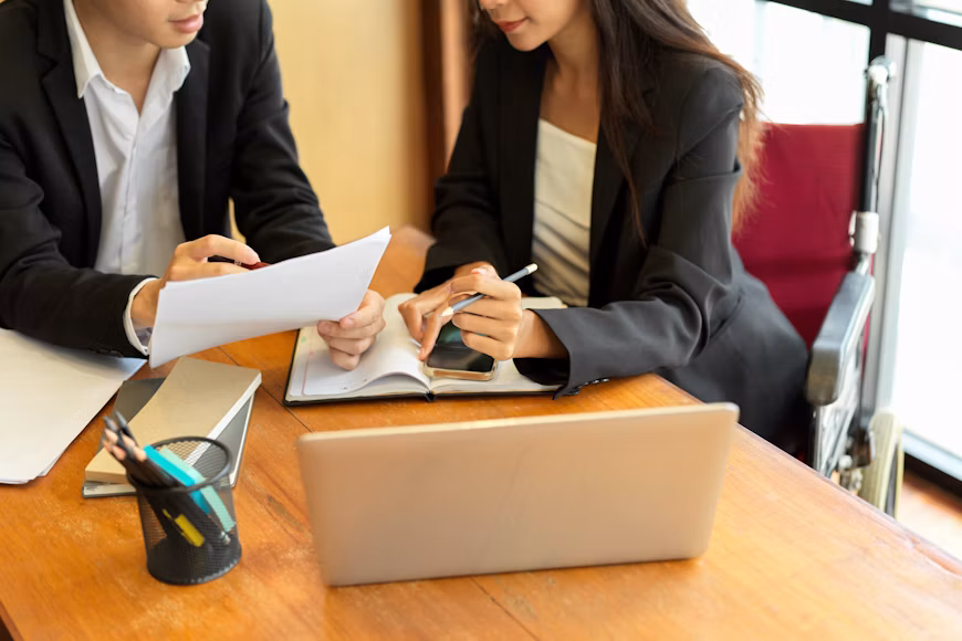 A business lawyer in Pasadena sits with a woman at a table with a laptop, documents, and a smartphone, with a wheelchair visible behind her.