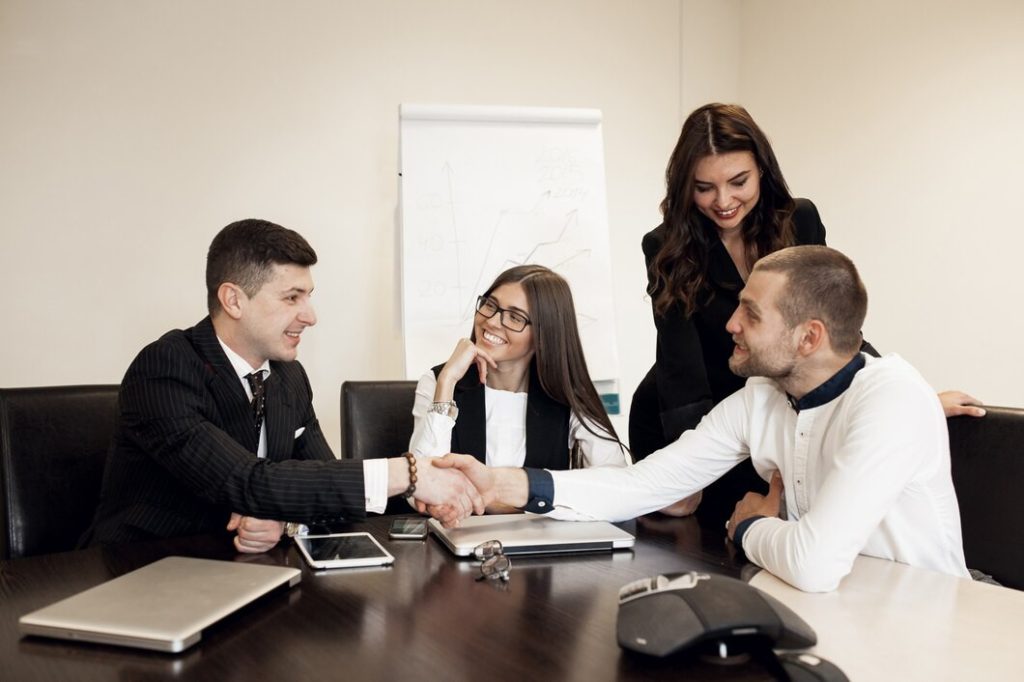 Two businessmen shake hands across a conference table while a business lawyer in San Pedro and another colleague look on.