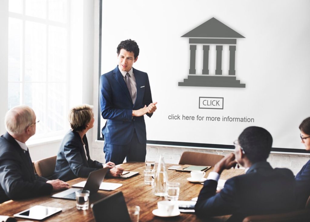 A business lawyer in Santa Ana presents to a group at a conference table, with a projection screen displaying a building icon and the text “CLICK” and “click here for more information.”
