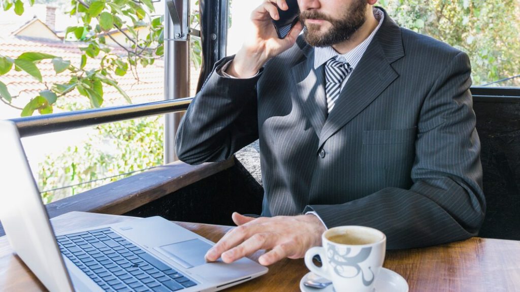 A Sherman Oaks business lawyer talks on a phone while using a laptop at an outdoor table with a cup of coffee.
