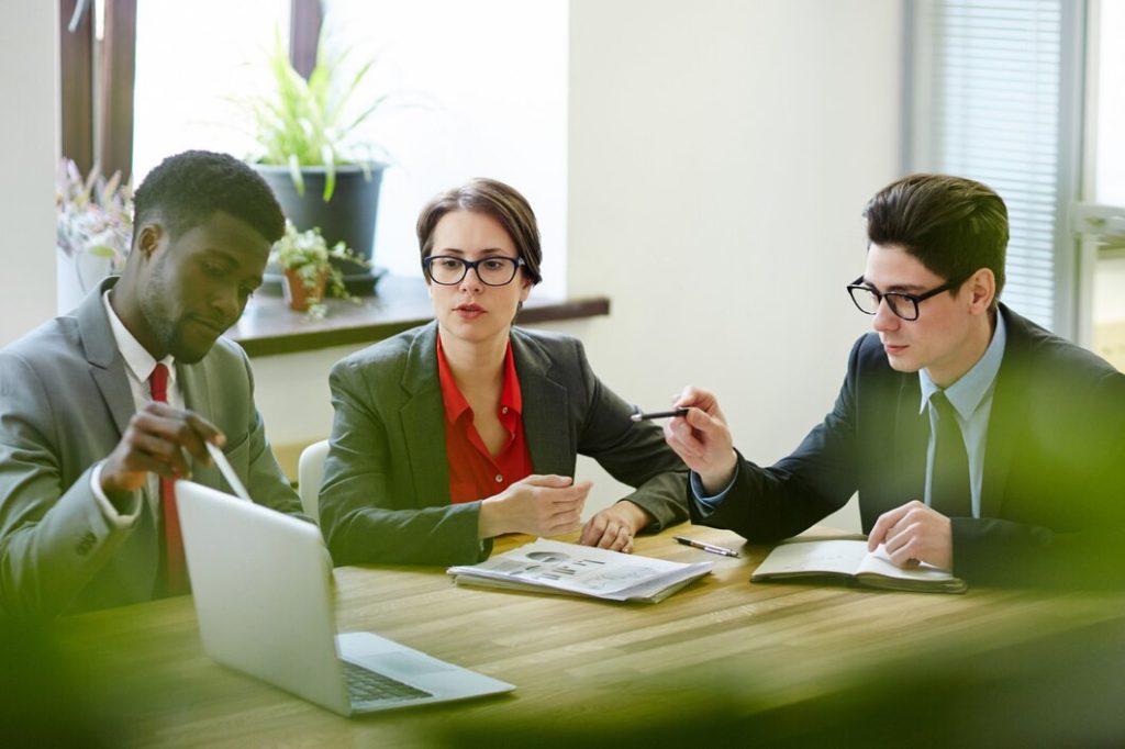 Three business lawyers in Temple City sit at a wooden table in an office, discussing documents and a laptop.