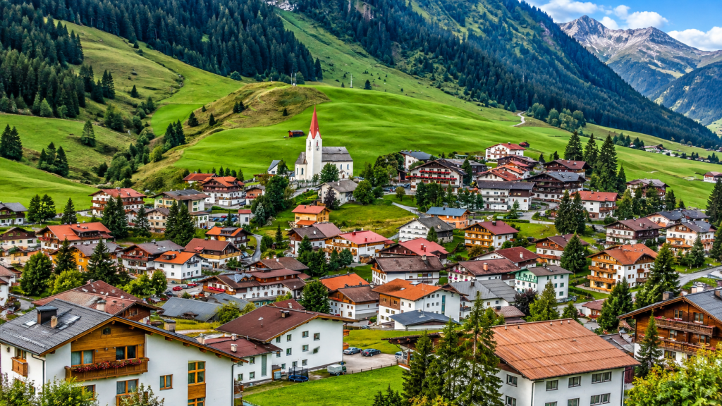A valley village with traditional houses and a white church with a red spire, nestled beneath green, forested mountains.