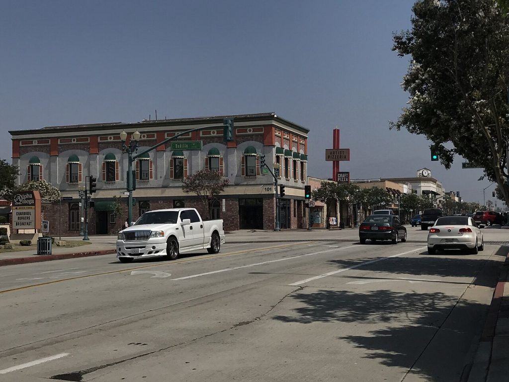 A street intersection in Covina near a business lawyer’s office, featuring a white pickup, a dark sedan, and a white sedan with multi-story buildings and an Artist Pizzeria sign in the background.