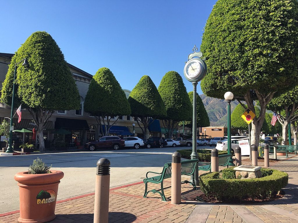 A sidewalk scene with manicured trees, a street clock, and benches outside storefronts including a business lawyer in Glendora, with cars parked along the street.