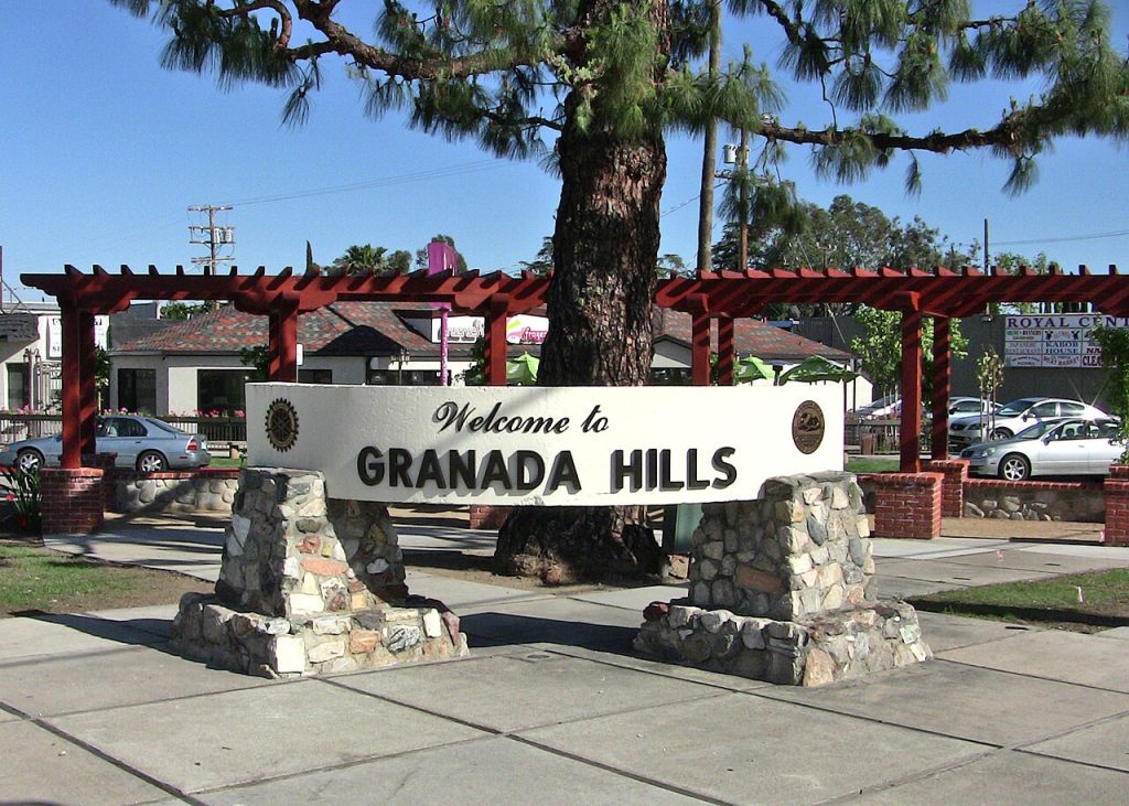 “Welcome to GRANADA HILLS” monument sign near a Business Lawyer in Granada Hills, supported by two stone pillars in front of a pine tree and a red wooden trellis.