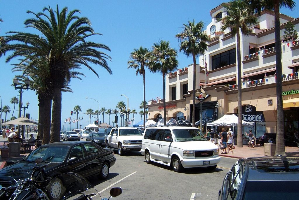 A street scene in a sunny, palm-lined Huntington Beach town with vehicles driving past local shops and a business lawyer office.