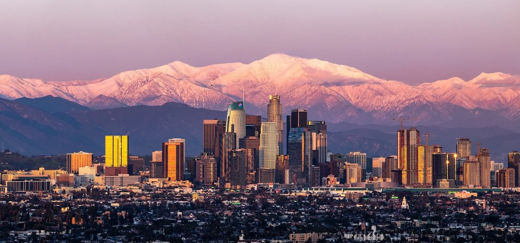 The Los Angeles skyline at sunset with snow-capped mountains in the background, viewable from a business lawyer in DTLA.