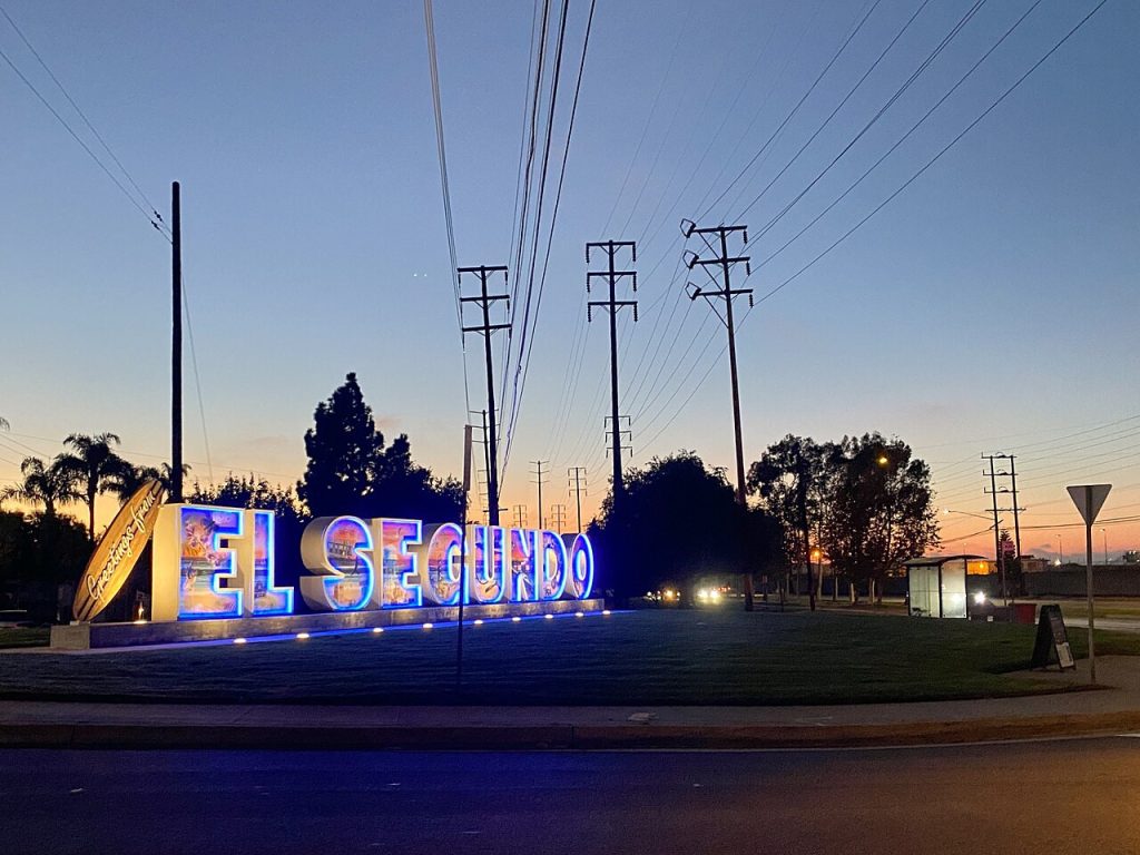The illuminated “EL SEGUNDO” sign at dusk, near a surfboard sculpture and utility poles, serving a community that relies on a local business lawyer in El Segundo.