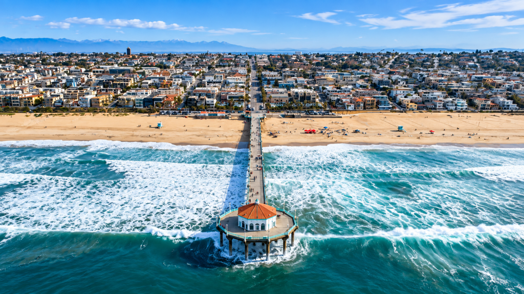 Aerial view of the Manhattan Beach pier near the office of a business lawyer serving South Bay, extending from the sandy beach into the ocean with the town and mountains in the background.