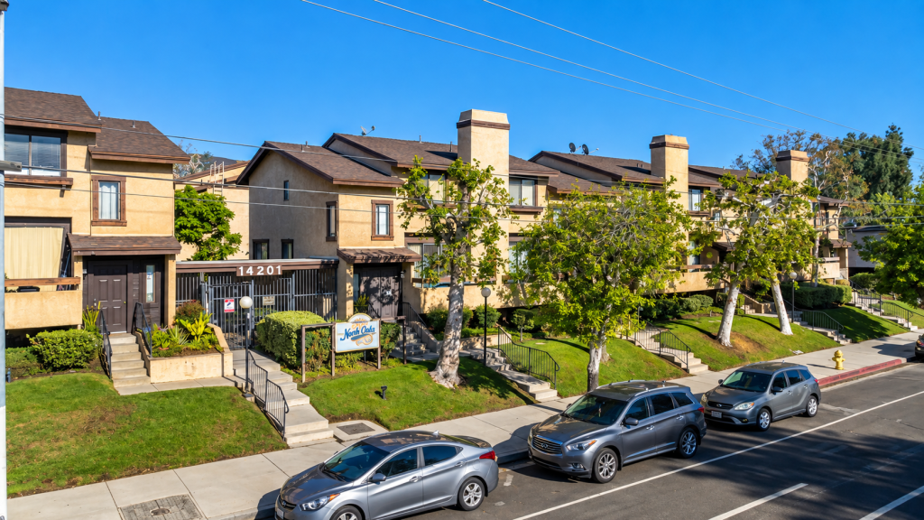 Multi-story tan townhome complex with brown roofs and a “North Oaks” sign at 14201, seen from the street where a business lawyer in Sylmar might visit, with parked cars in the foreground.