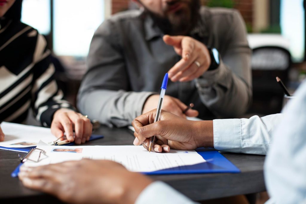 A person signs a document on a clipboard to meet corporate compliance requirements during a meeting while others look on.