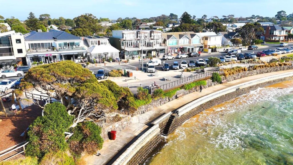 Aerial view of a coastal street in Brentwood, lined with shops, a parking lot, and a stone seawall bordering the ocean.