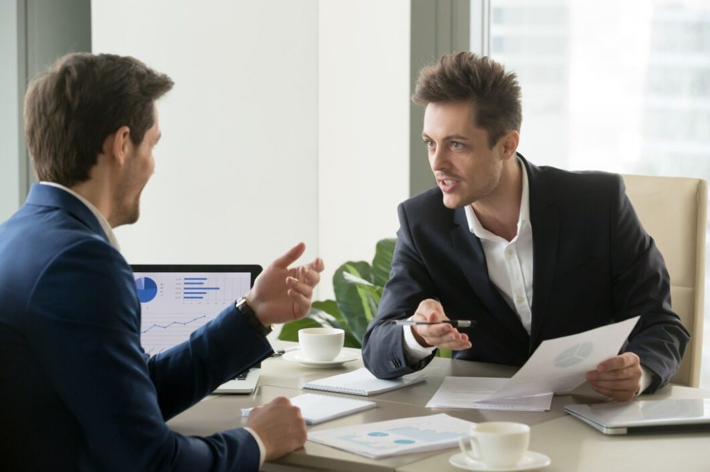 Two businessmen in suits having a discussion across a desk with a business disputes lawyer, featuring documents and a laptop showing a business chart.