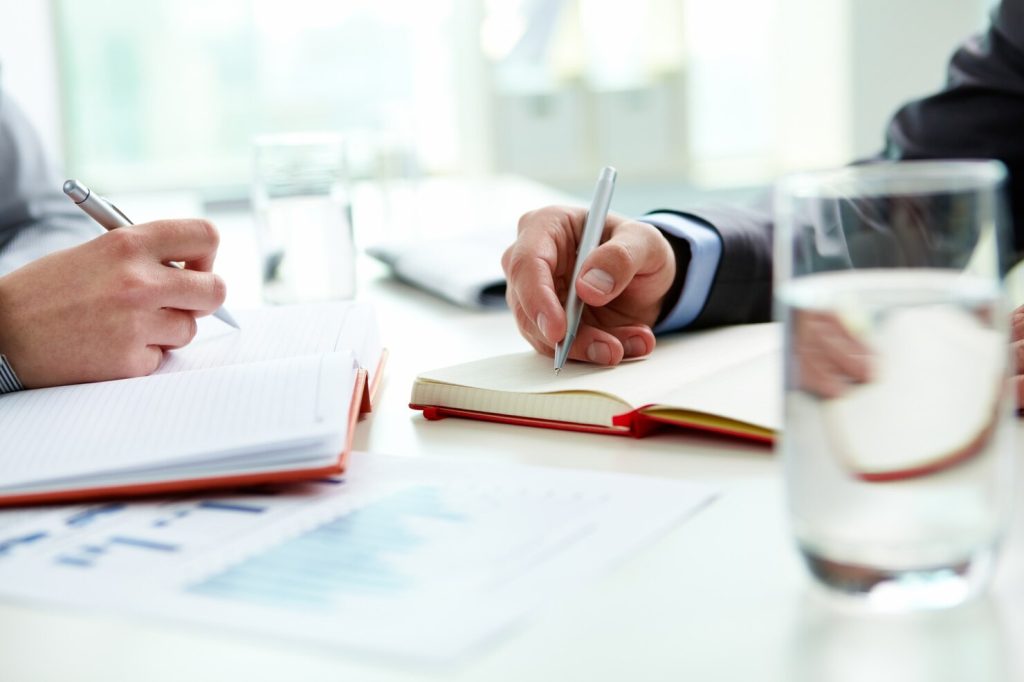 Two people writing in notebooks at a table with documents and glasses of water while reviewing corporate compliance requirements.