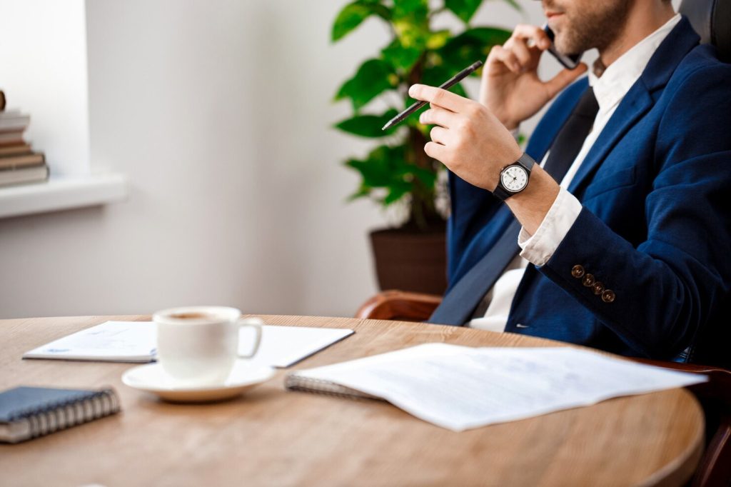 A business lawyer in Carson sits at a wooden table in a blue suit, talking on a smartphone and holding a pen, with documents and a coffee cup in front of him.