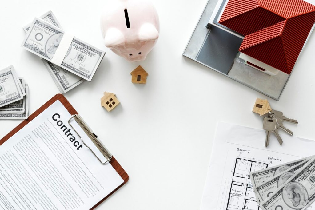 A real estate law attorney’s contract on a clipboard, a piggy bank, bundles of cash, a model house, house keys, and a floor plan on a white surface.