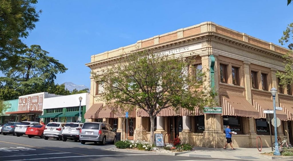 Street view of a two-story tan brick building housing a business lawyer in Claremont, next to a row of smaller shops and a parked line of cars.