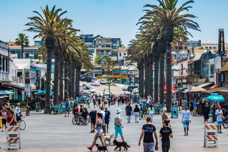Pedestrians walk down a wide plaza in Hermosa Beach lined with palm trees, shops, and buildings, near the office of a business lawyer.