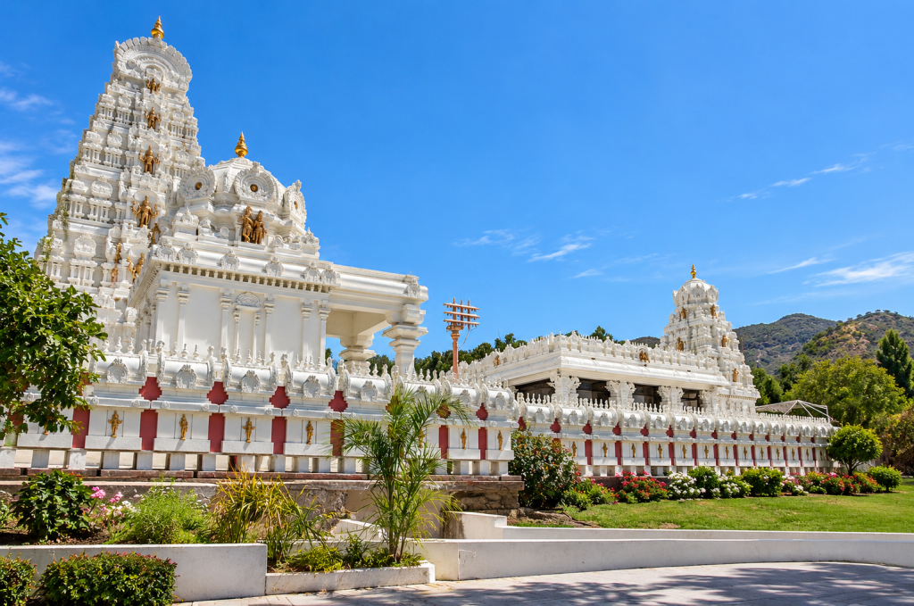 A white Hindu temple with ornate spires and red accents stands near a Malibu business lawyer office, set against a bright blue sky and grassy landscape.