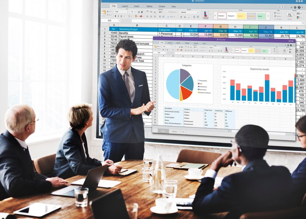 A corporate law attorney in Los Angeles presents a spreadsheet and charts on a large screen to colleagues in a boardroom.