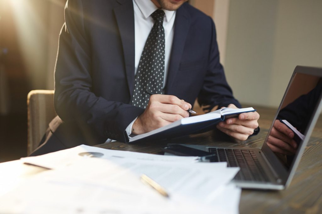 A Hollywood business lawyer in a suit sits at a desk, writing in a notebook next to a laptop and documents.