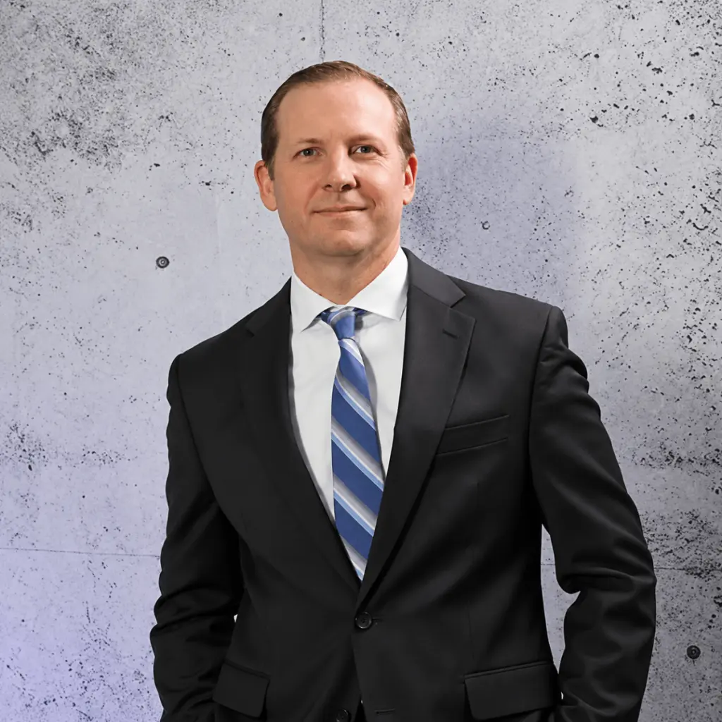 A man from Lurie & Associates Business Litigation Lawyers in a black suit, white dress shirt, and blue striped tie standing against a concrete wall.